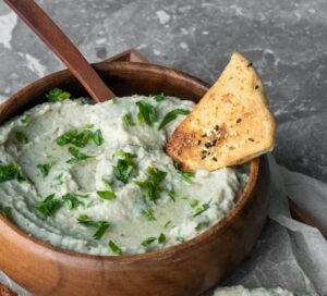 Baba Ghanouj (Ganuosh) in a wooden bowl with bright green spices and a fresh baked chip
